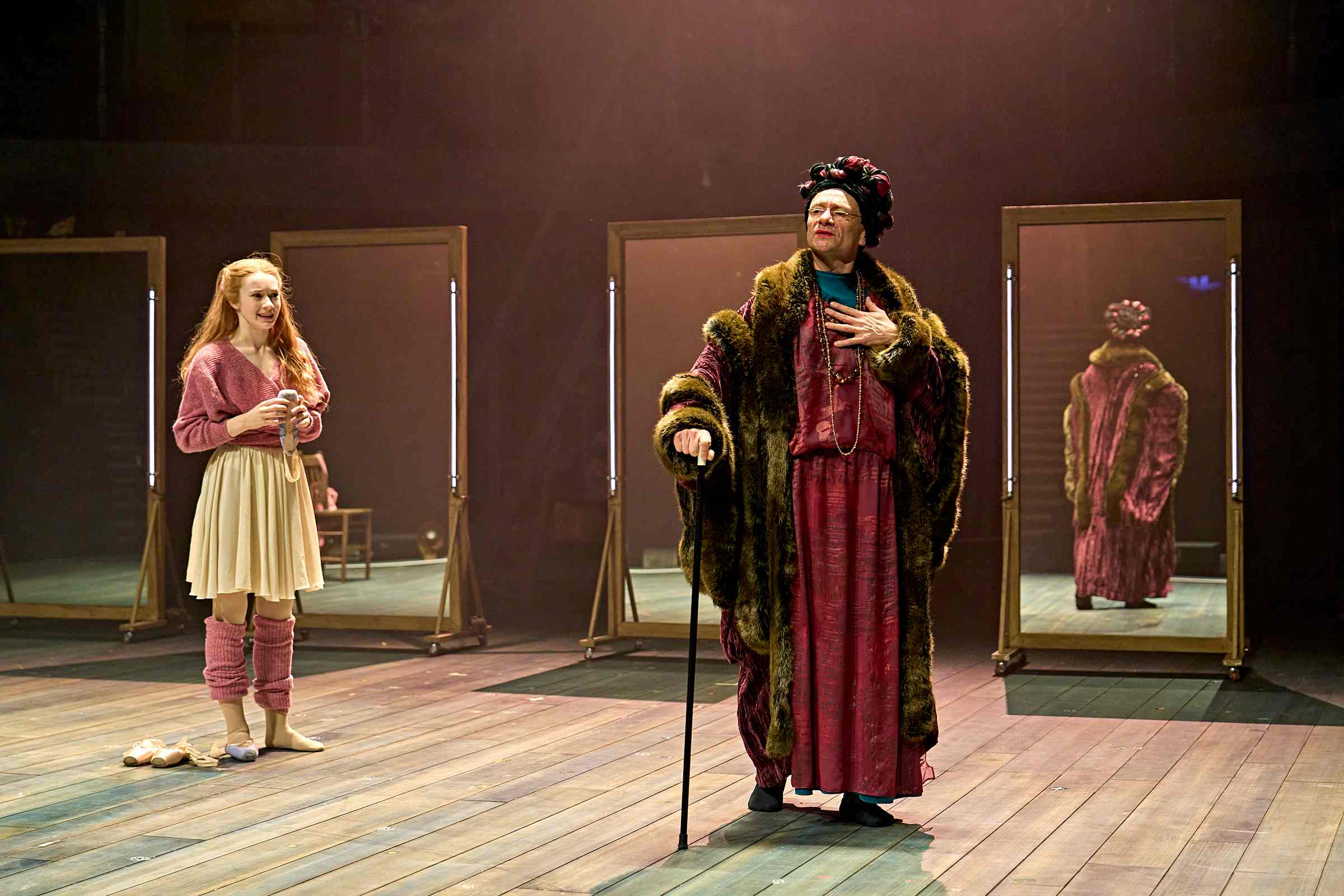 A scene from the stage production Ballet Shoes, showing a young woman in ballet attire—leg warmers, leotard, and skirt—standing barefoot and holding a pair of pointe shoes. Beside her, an older character in a dramatic fur-trimmed robe and headdress holds a cane and gestures grandly, with mirrors behind reflecting their theatrical costumes.